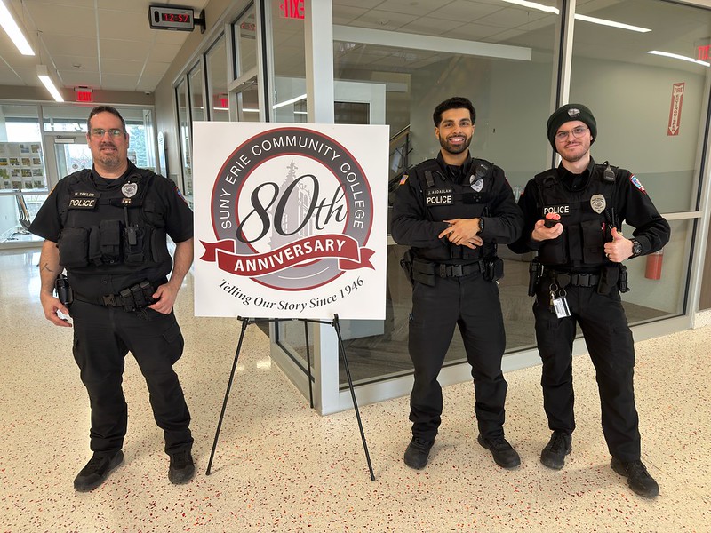 Three campus police officers stand beside an 80th anniversary sign for SUNY Erie Community College inside the STEM Building at North Campus.