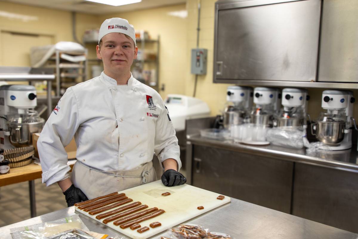 A culinary arts student stands in a professional teaching kitchen, preparing confections at a workstation surrounded by commercial mixers and food production equipment. The student wears a SUNY Erie chef’s uniform, gloves, and hat.