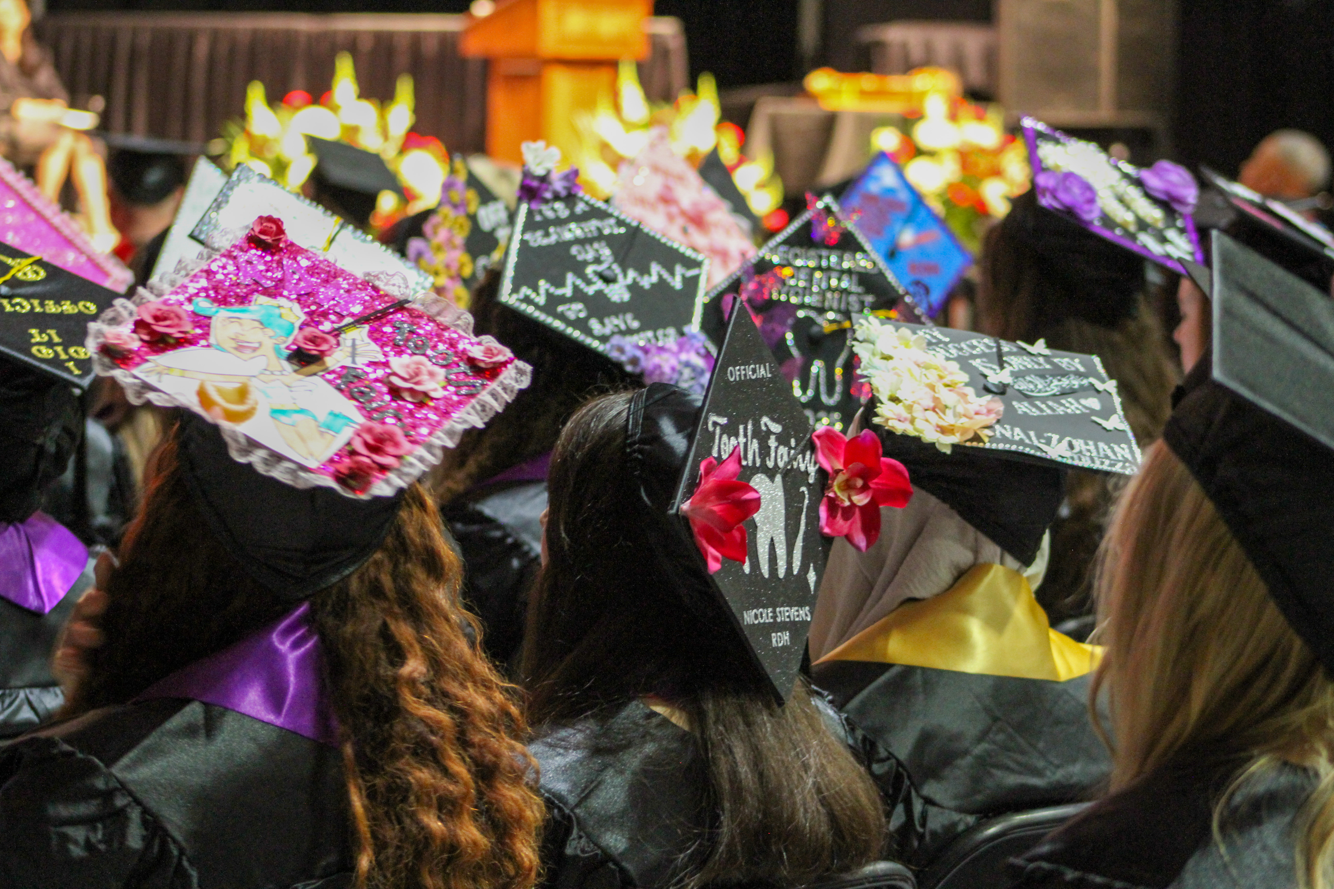 Graduates sit together at a commencement ceremony, wearing decorated caps adorned with colorful designs, messages, and embellishments as they face the stage.