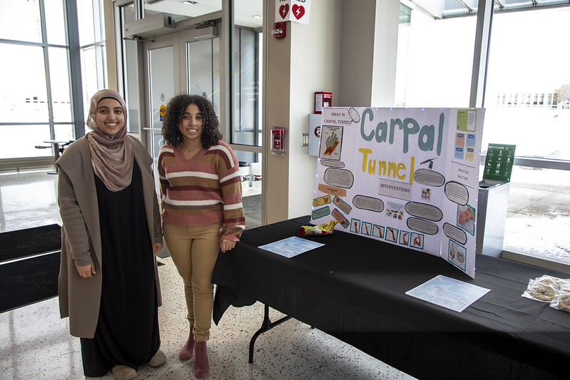 Two students stand beside a display table with a presentation board about carpal tunnel syndrome and treatment interventions during a student project showcase at SUNY Erie.