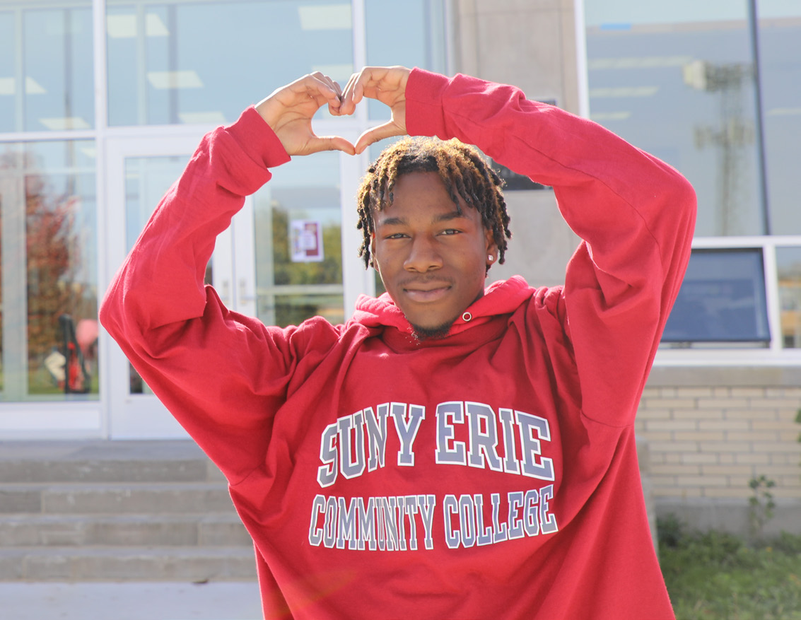 A student wearing a red “SUNY Erie Community College” hoodie stands outside a the north campus student center, smiling softly and forming a heart shape with their hands above their head.