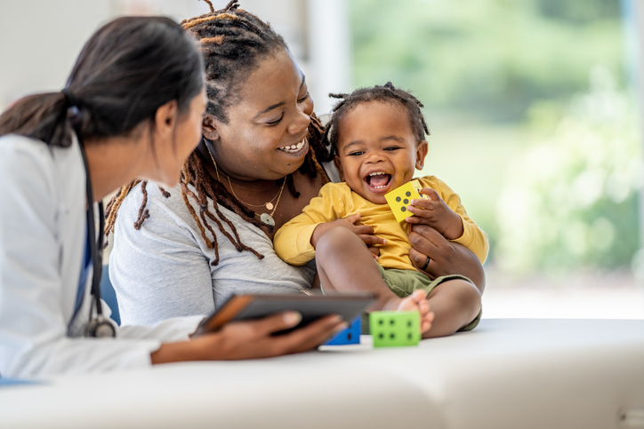 A young Mother, of African decent, brings in her toddler for a check-up. She is holding the little girl gently on the exam table as she talks with the doctor who is holding out a tablet with test results on it.